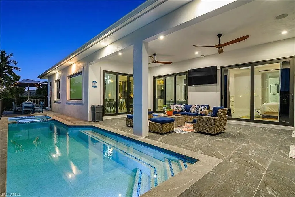 Covered lanai at dusk with a sofa and wicker seating, a wall-mounted television, ceiling fans and the lit pool and spa beside it.