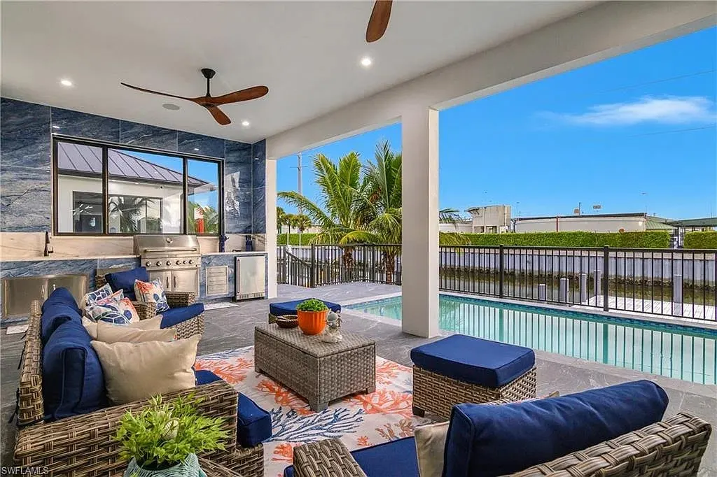 Covered lanai with an outdoor kitchen set against blue stone-look tile, a grill, a sink and wicker seating overlooking the pool and canal.
