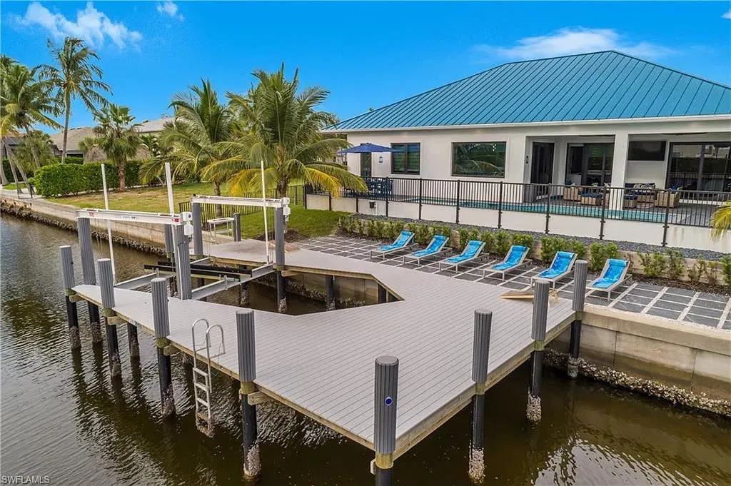 Waterfront deck and private boat dock with a boat lift, lounge chairs on a paver terrace and the blue-roofed home beyond.