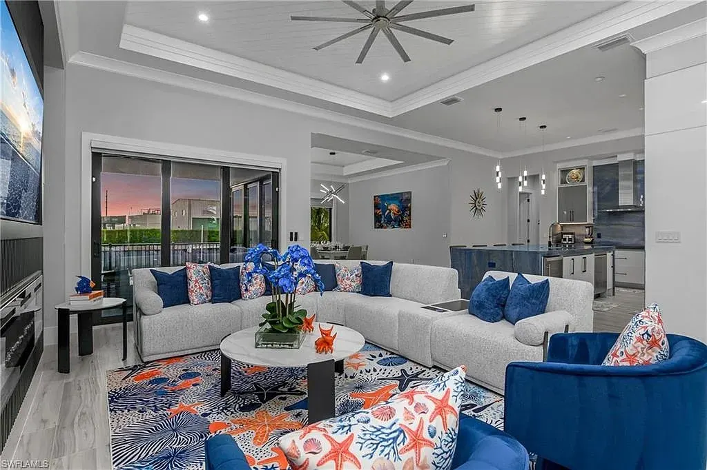 Living room with a gray sectional, blue accent chairs, a tray ceiling with white planks and sliding glass doors to the waterfront, with the kitchen beyond.