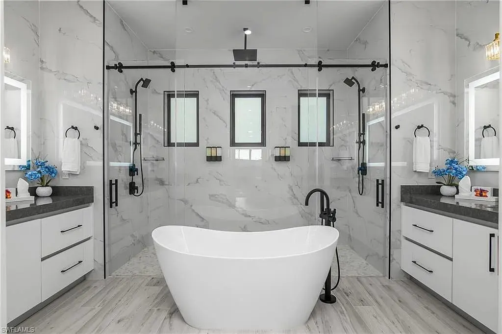 Bath with a freestanding tub between two vanities and a glass-enclosed marble-look shower.