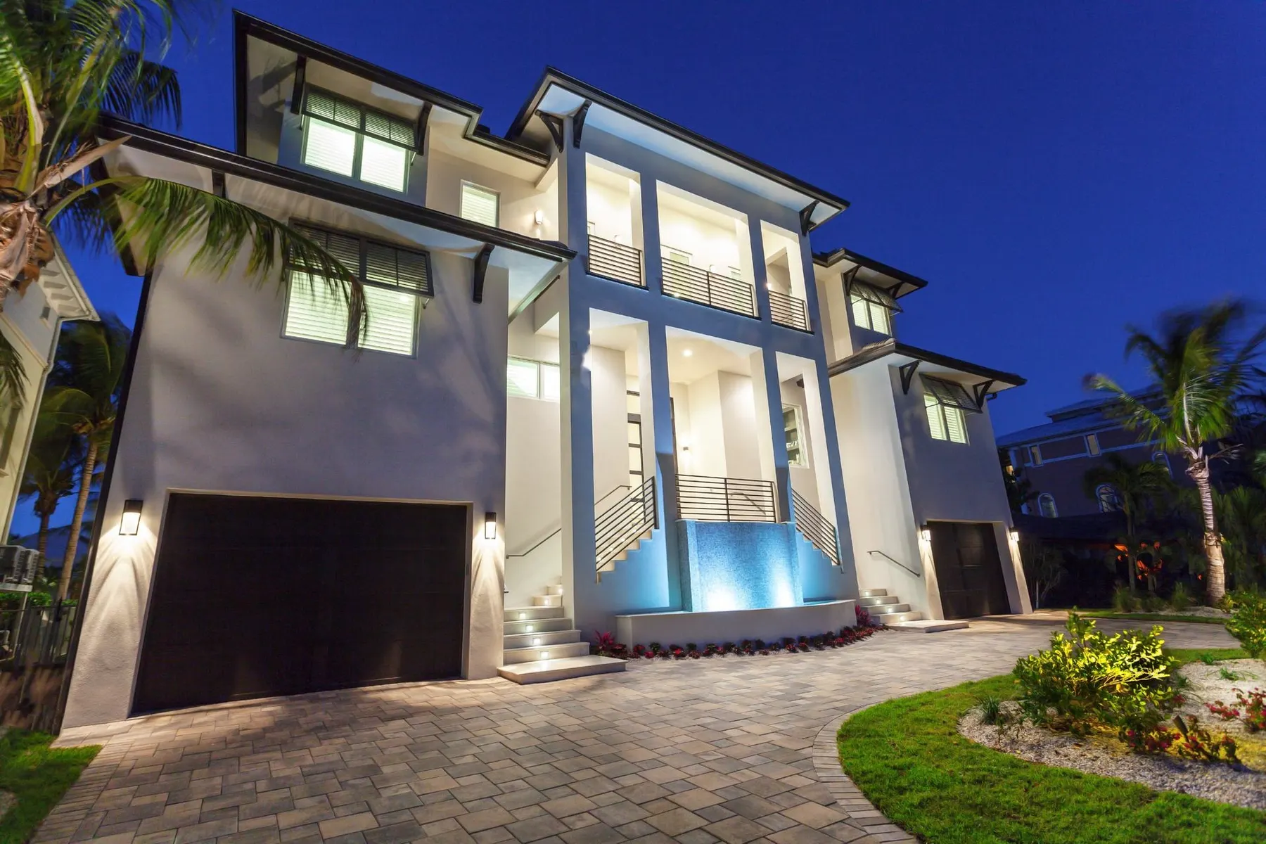 Front of the residence at dusk with lit entry columns, a glass-tile water feature and paver drive.