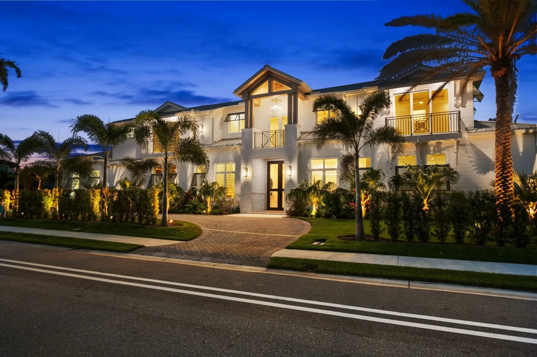 Front of the Moorings Harbor Drive Residence at dusk, a two-story white home with a gabled entry, a metal roof and a curved paver driveway lit by landscape lighting.