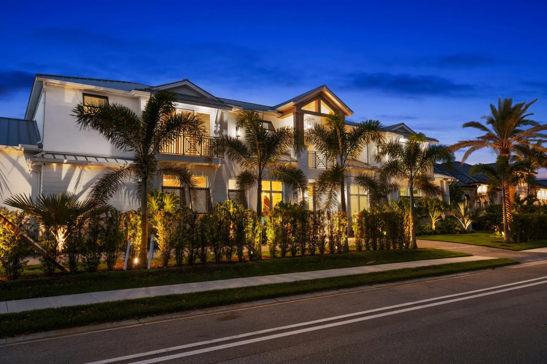 Front of the home at dusk seen from the street, with uplit palms, balconies with crossed railings and a tall hedge along the sidewalk.