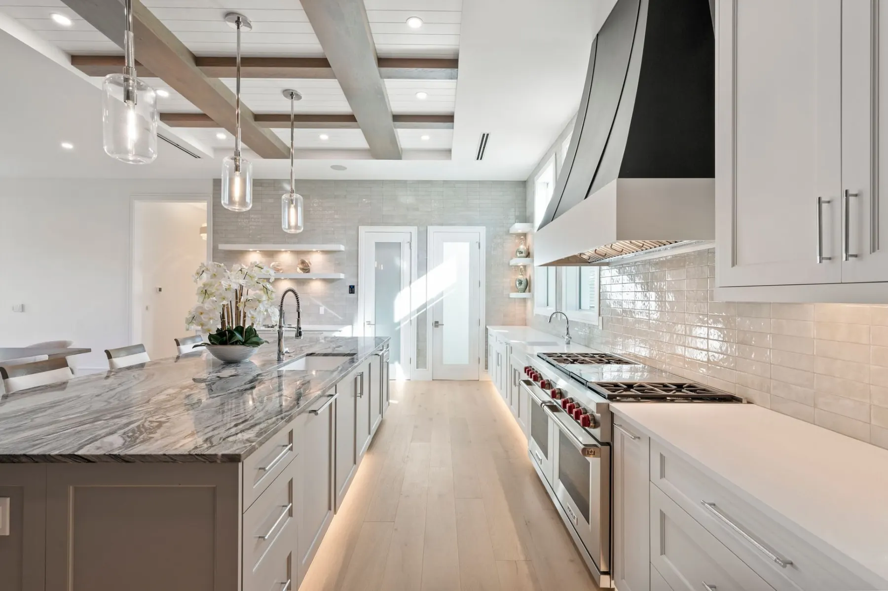 Kitchen with a long island, a black range hood, a gas range and white cabinets under a coffered ceiling.