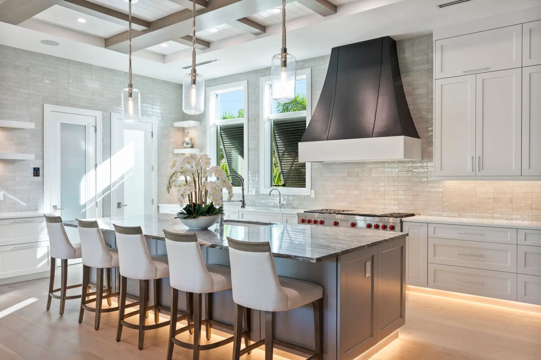 Kitchen island with counter stools and under-counter lighting, with a black hood and tall windows.