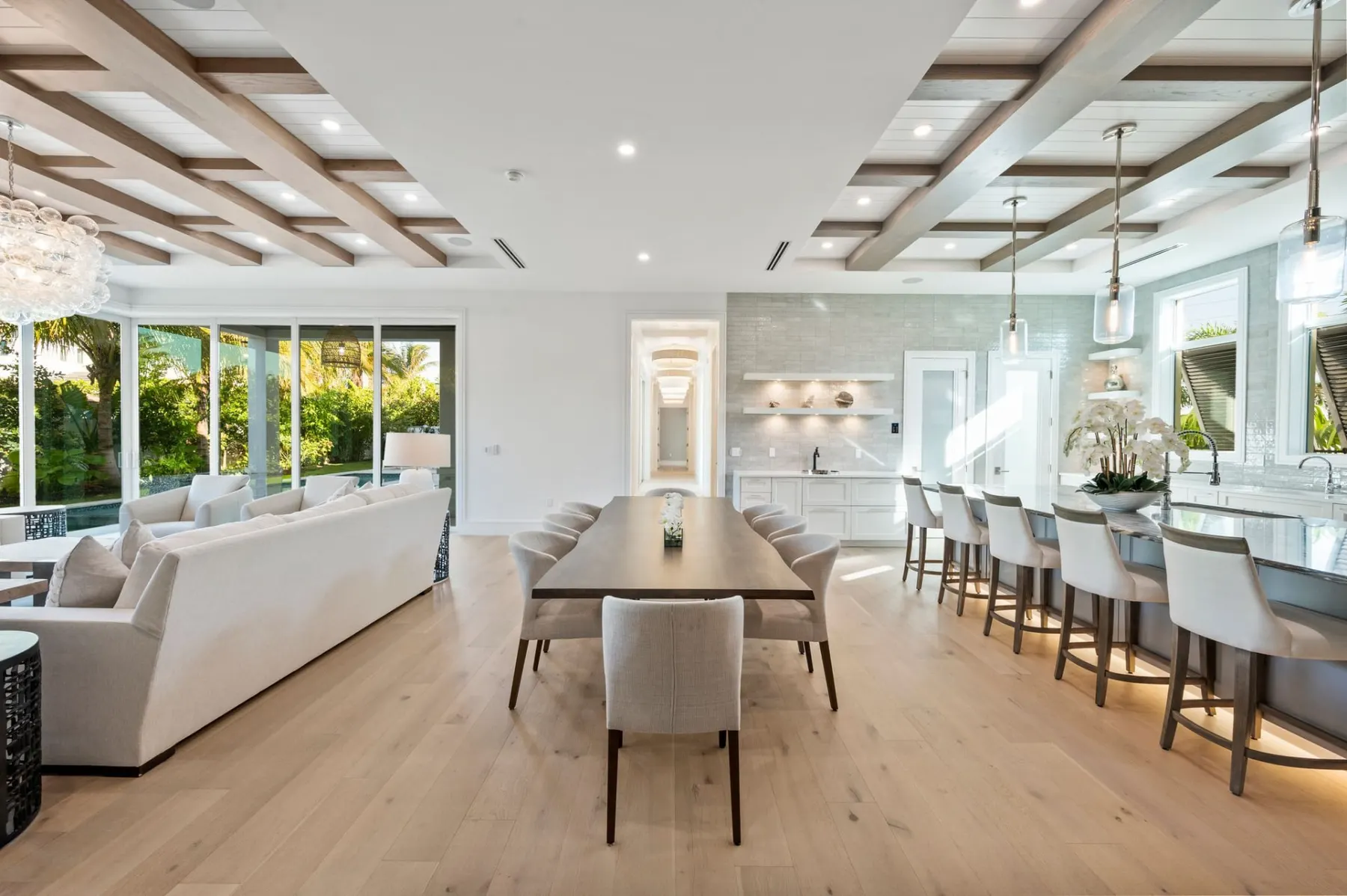 Dining table with a long kitchen island and counter stools, and the living room beyond, under coffered beams.
