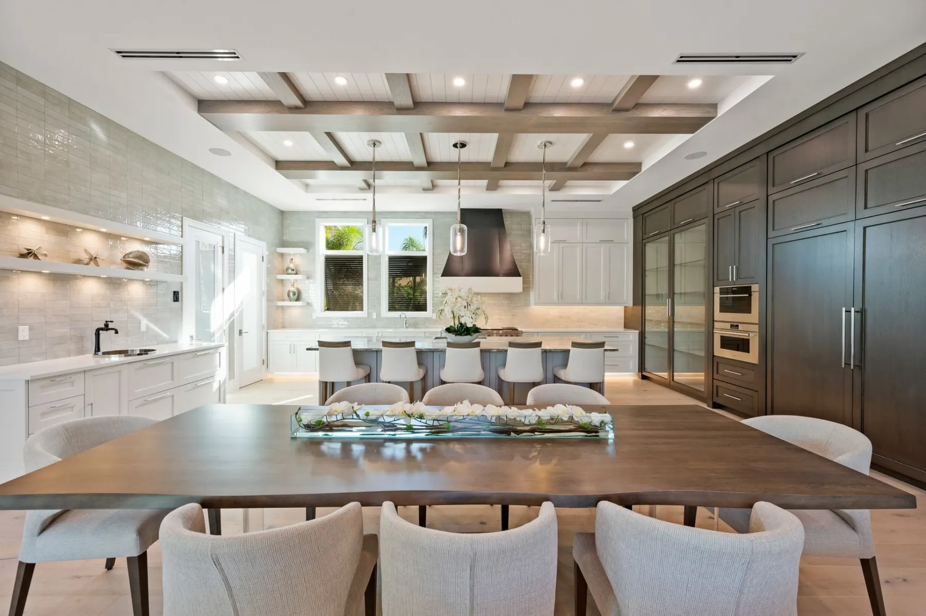 Dining table and upholstered chairs with the kitchen, dark cabinets and a black range hood beyond.