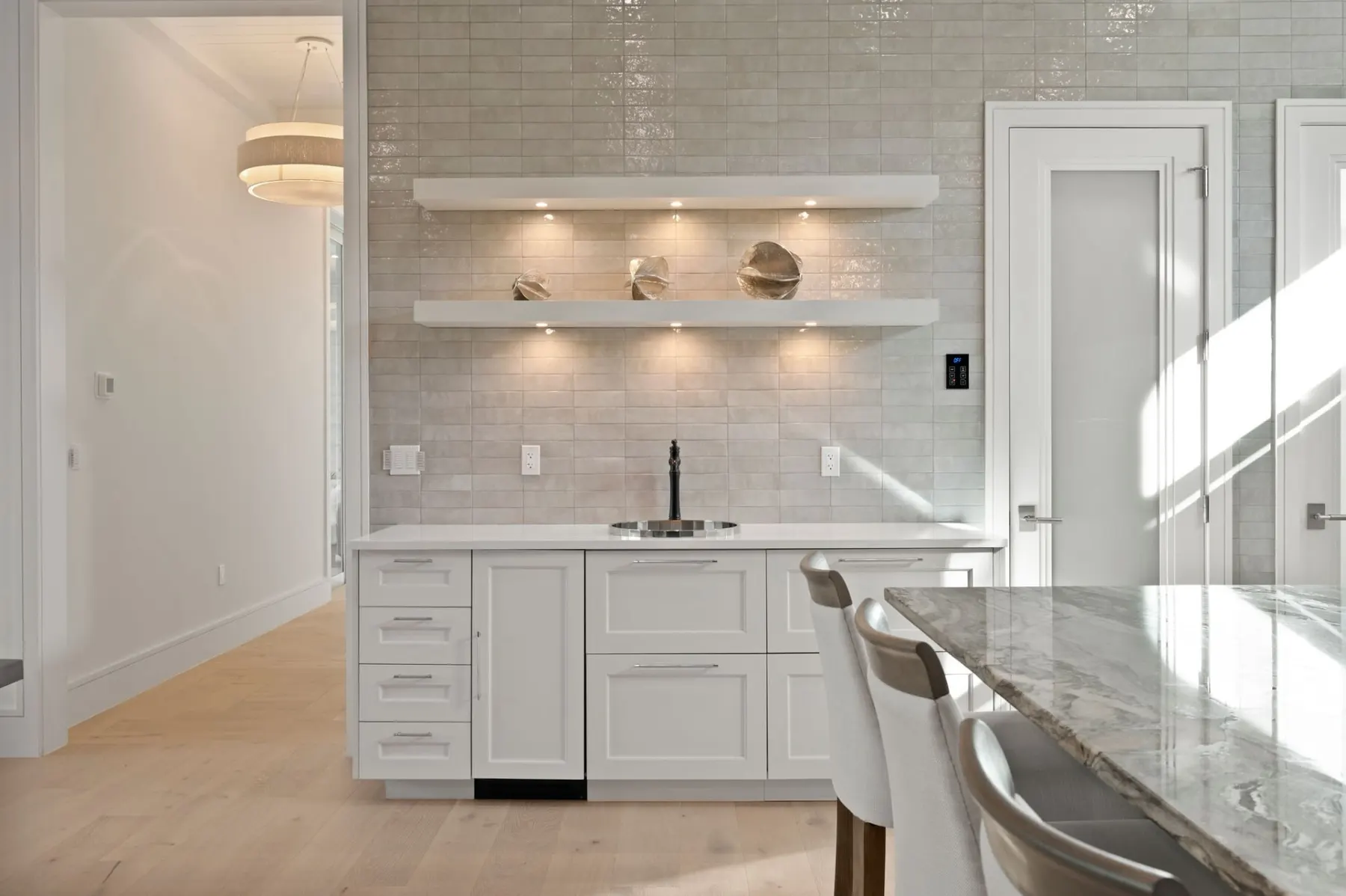 Wet bar with glazed tile, lit open shelves, a small sink and white cabinets beside the kitchen.