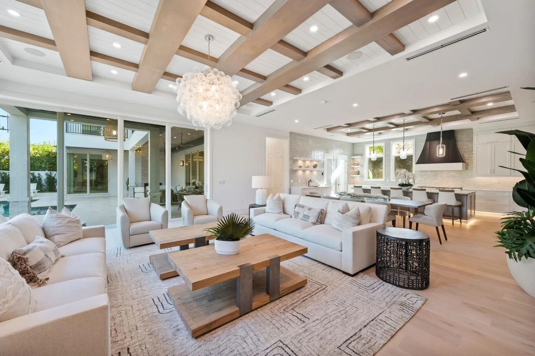 Open living and dining space with white sofas, a patterned rug and a bubble chandelier under a coffered beam ceiling.