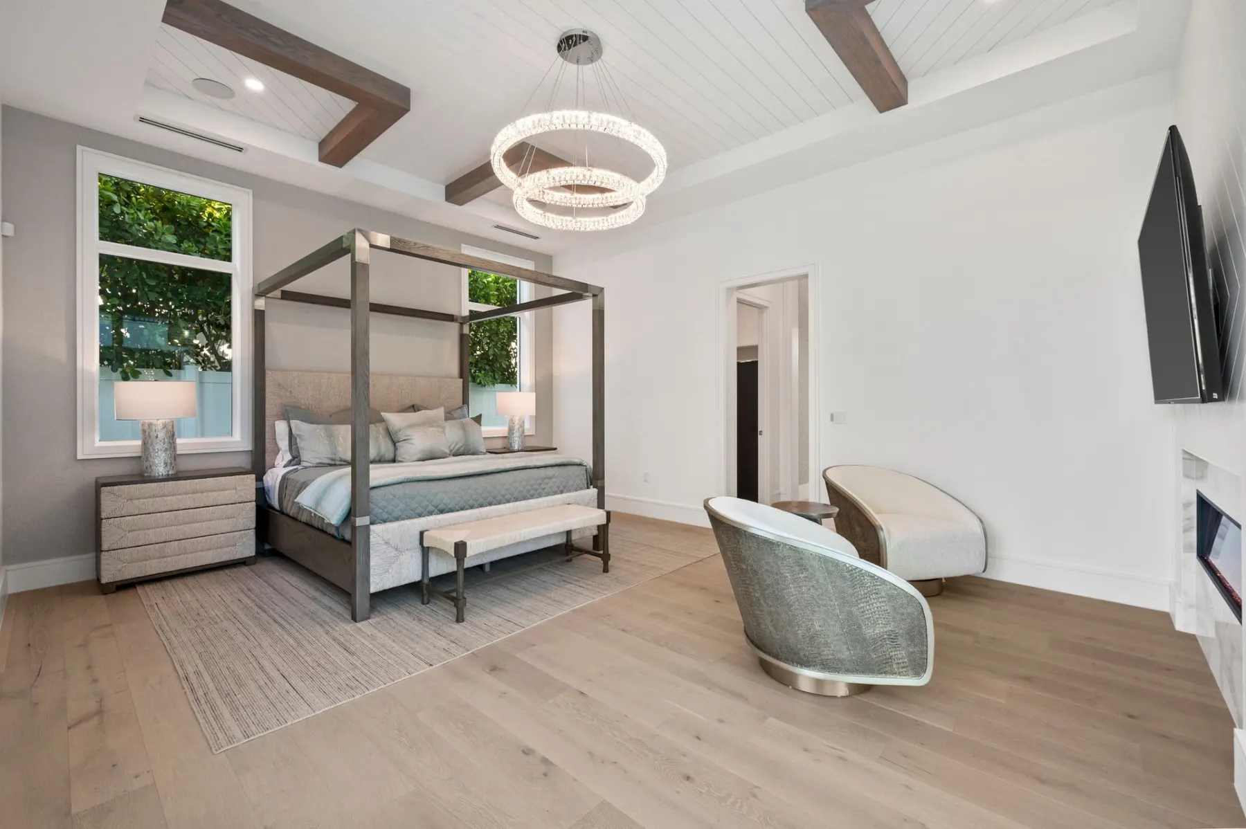 Primary bedroom with a canopy bed, a bench, an accent chair and a television, under a beamed tray ceiling.