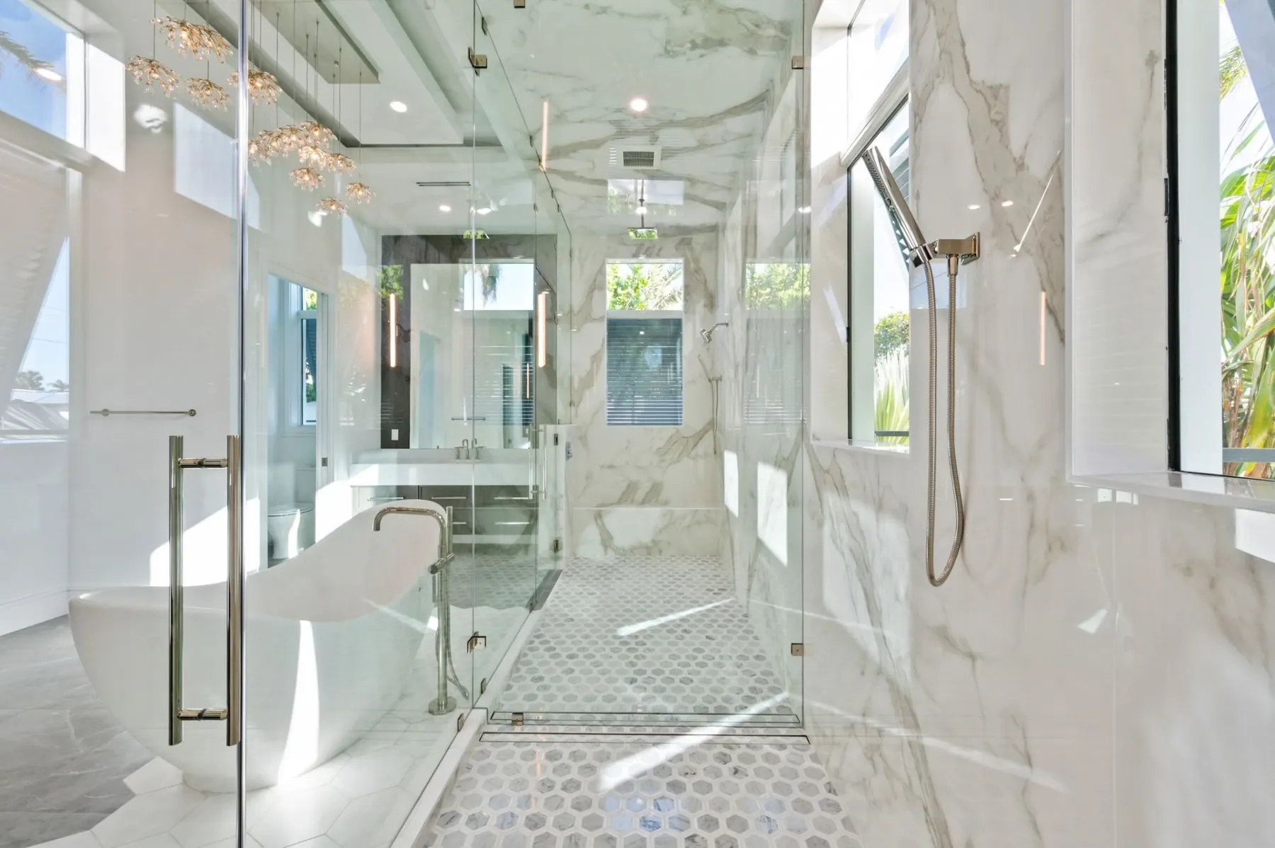 Primary shower with glass walls, white veined tile and a hand shower, with the tub beyond.