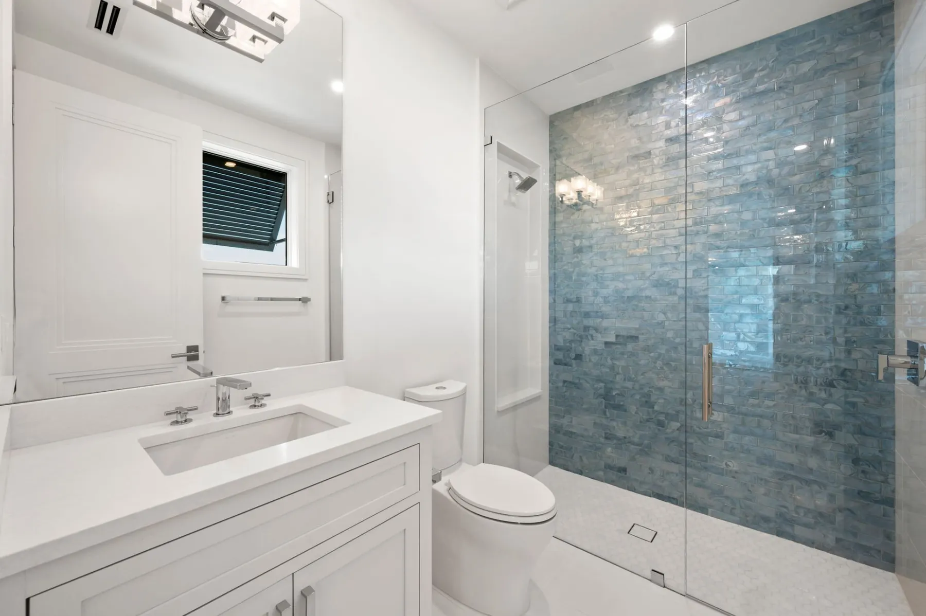 Bath with a blue textured tile shower, a white vanity and a window.
