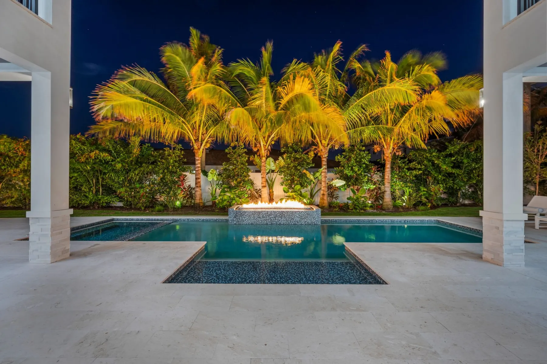 Pool and fire bowl at night with uplit palms, seen from the covered terrace.