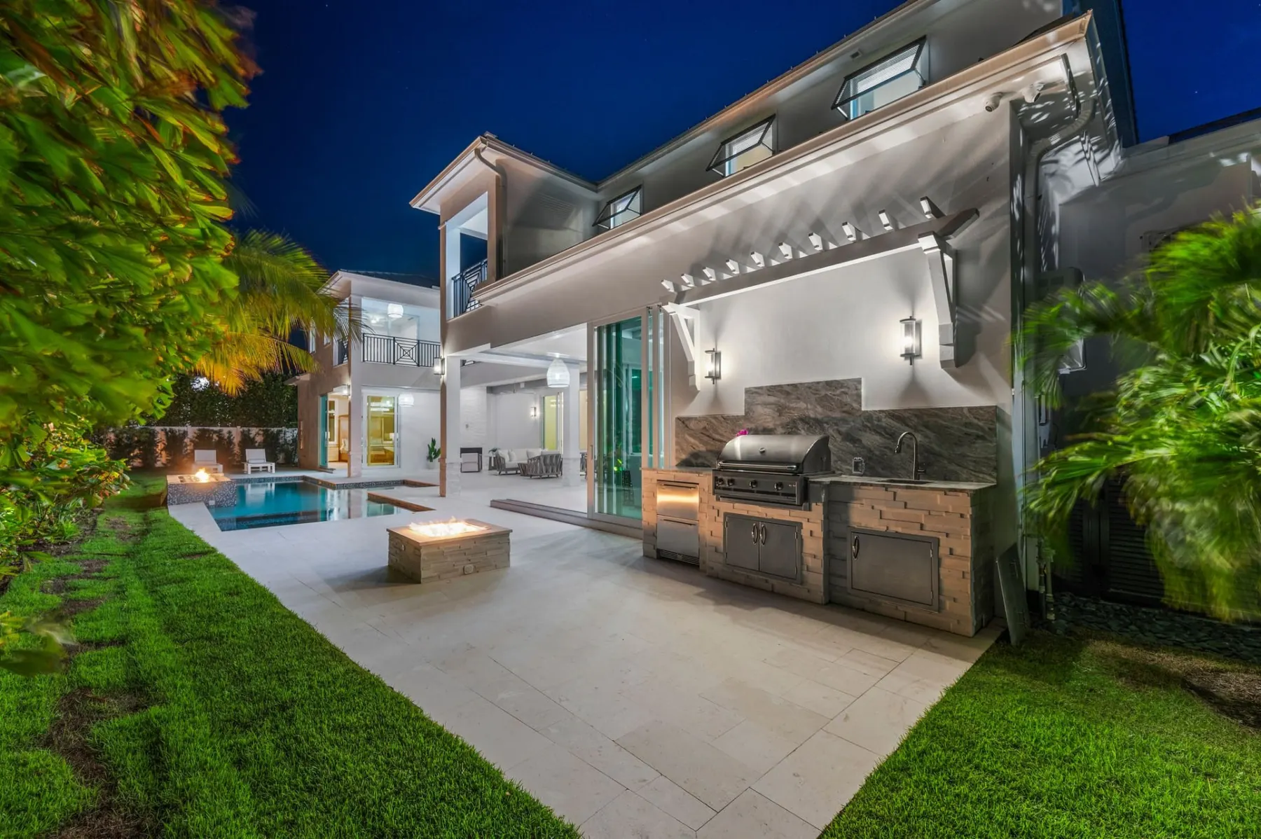 Outdoor kitchen and fire pit at dusk with the pool lit in the background.