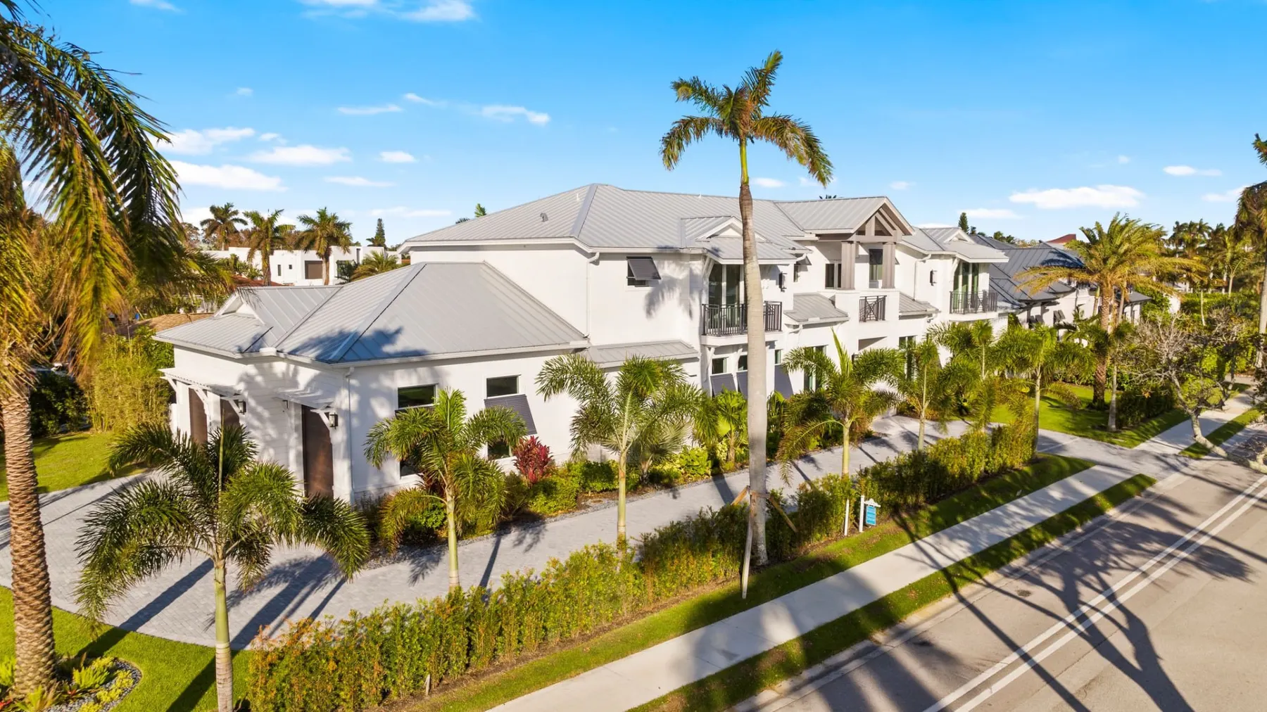 Aerial view from the street of the front and garage wing, with a standing-seam metal roof and palms along the paver driveway.