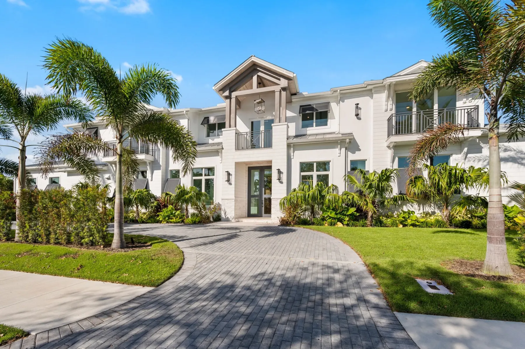 Front elevation by day with a tall gabled entry, white siding and stucco, bahama shutters and a curved paver driveway.