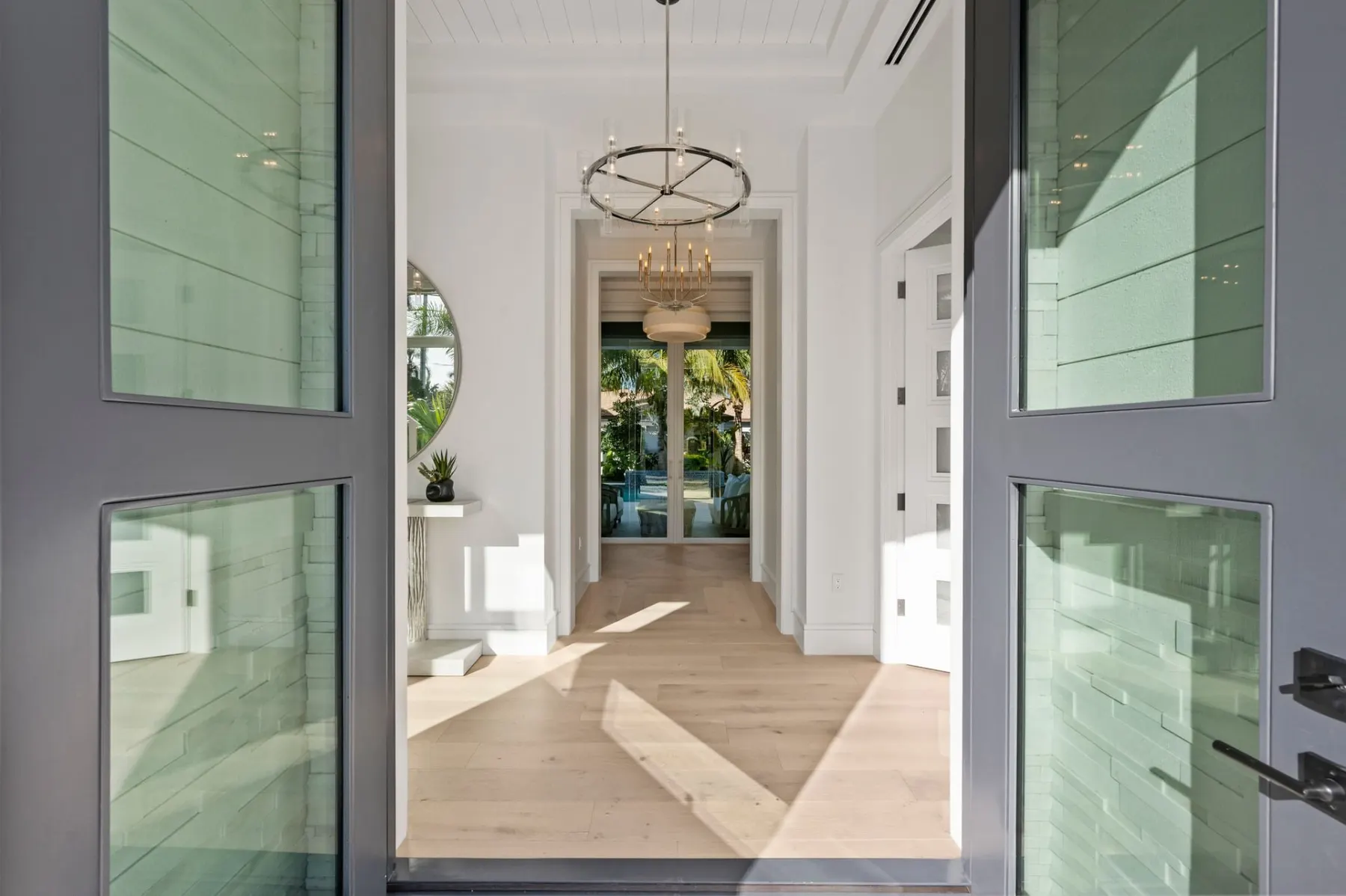 Open front doors looking down the foyer, with a ring chandelier, herringbone wood floors and the covered lanai beyond.
