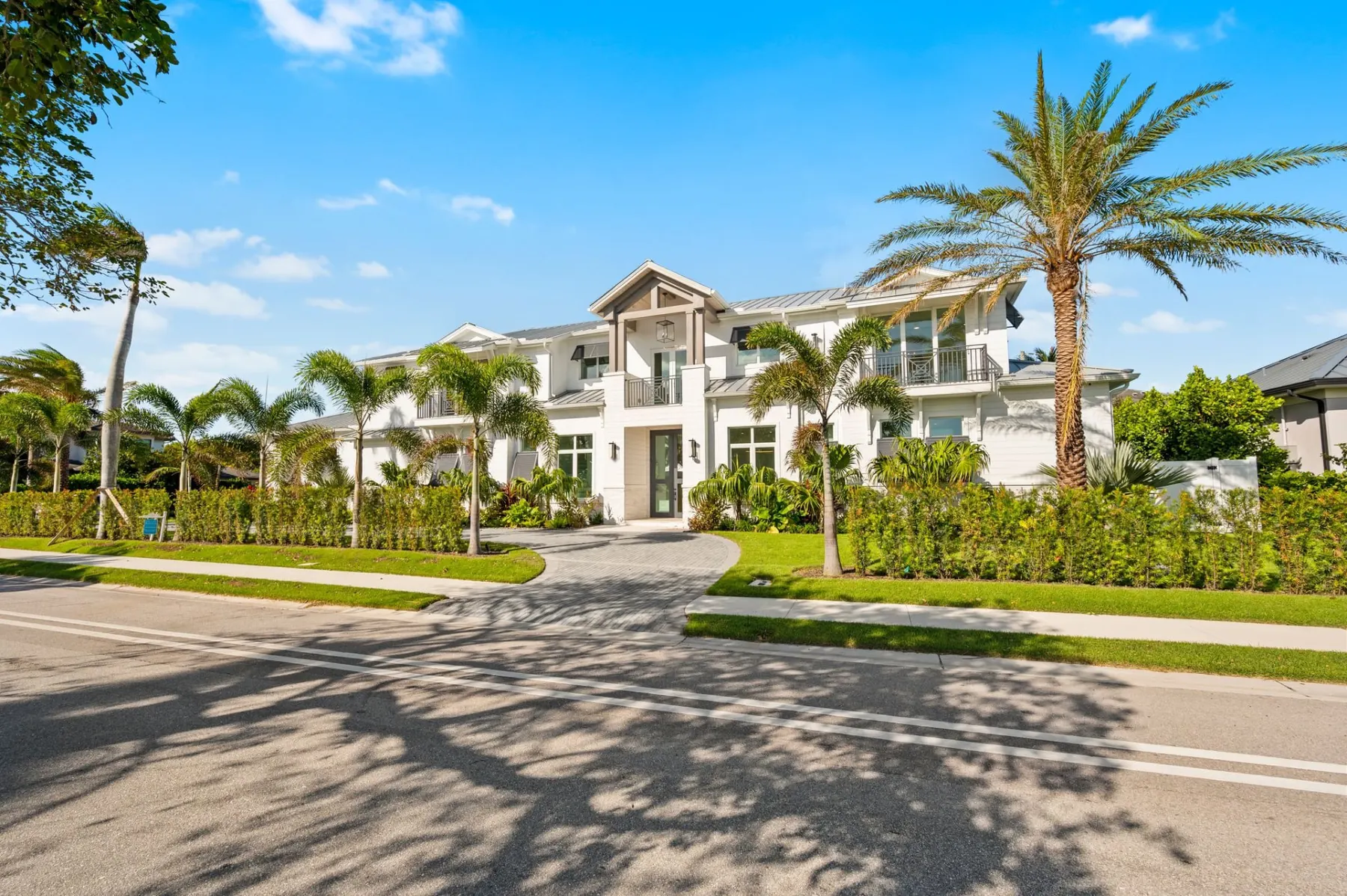 Front of the home from across the street, with a gabled entry, a balcony with crossed railings and palms in front of a hedge.