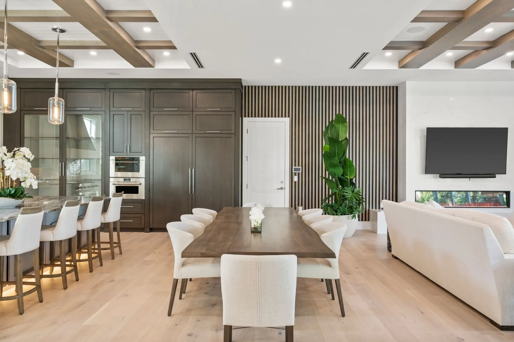 Dining table in front of a slatted feature wall, with dark kitchen cabinets and a white sofa facing a linear fireplace.