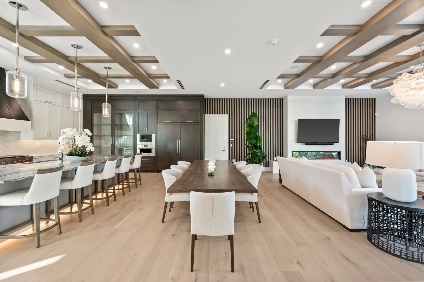 Wide view of the open kitchen, dining and living room, with a long island, a wood-beam ceiling and a bubble chandelier.