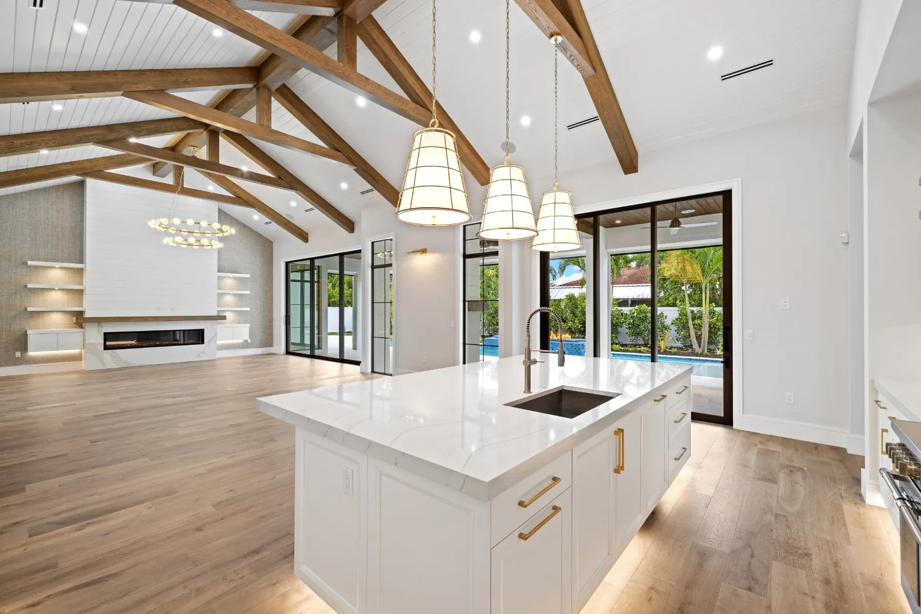 Open kitchen and great room with a large white island, a vaulted ceiling with timber trusses and a linear fireplace wall beyond.