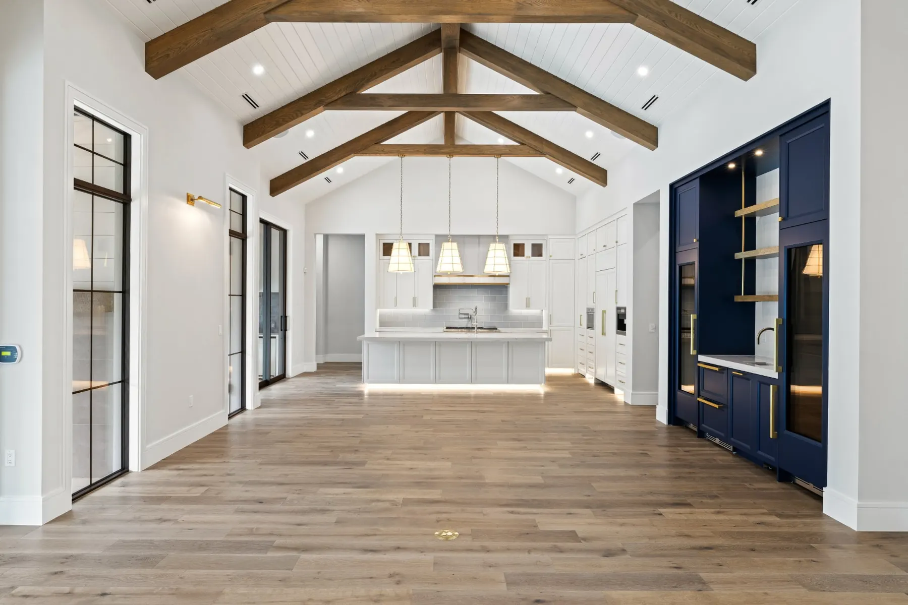 Great room looking toward the kitchen, with a navy bar cabinet with open shelves under timber trusses.
