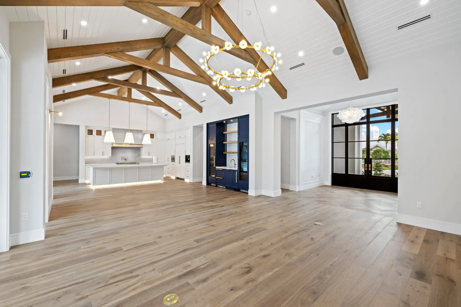 Wide view of the great room under timber trusses, with the kitchen at left and a navy bar and glass doors at right.