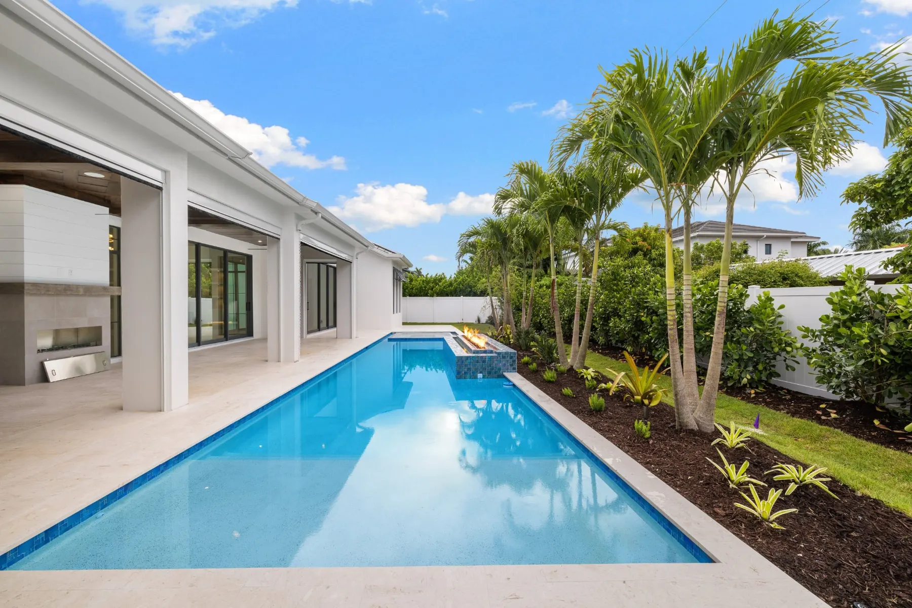 Long pool along the covered lanai in daylight, with a fire feature and a palm bed.