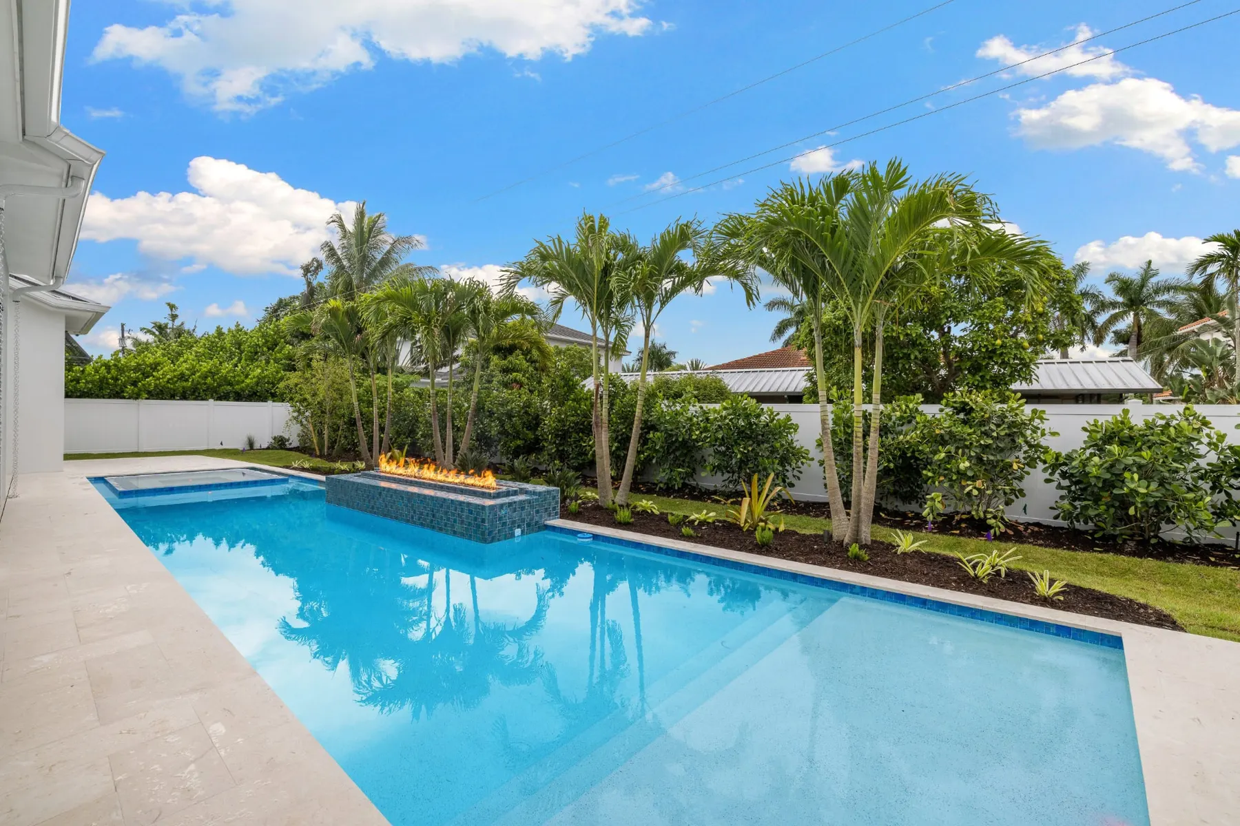 Pool and fire feature in daylight with palms and a white privacy fence.