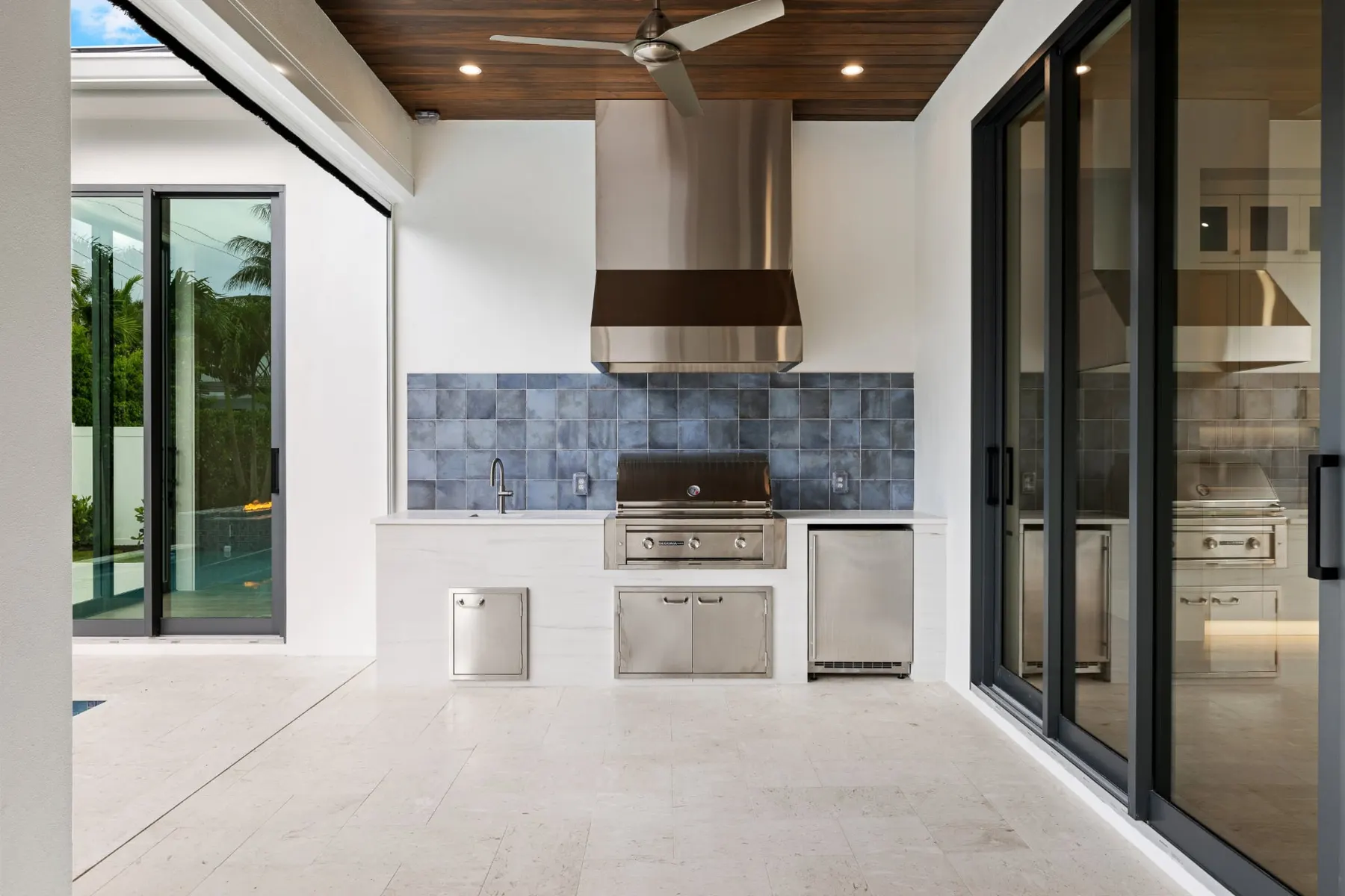 Outdoor kitchen with a stainless hood, grill, sink and beverage refrigerator against blue tile.