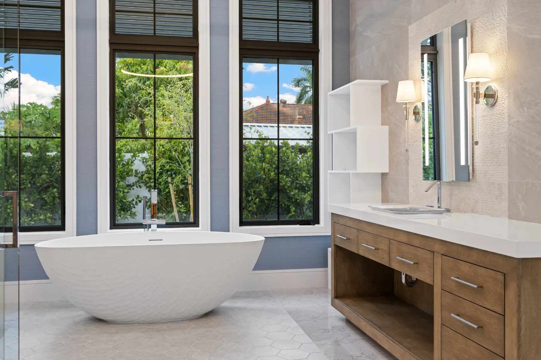Bath with a freestanding sculpted tub beneath three tall windows, beside a wood vanity with a lit mirror and sconces.