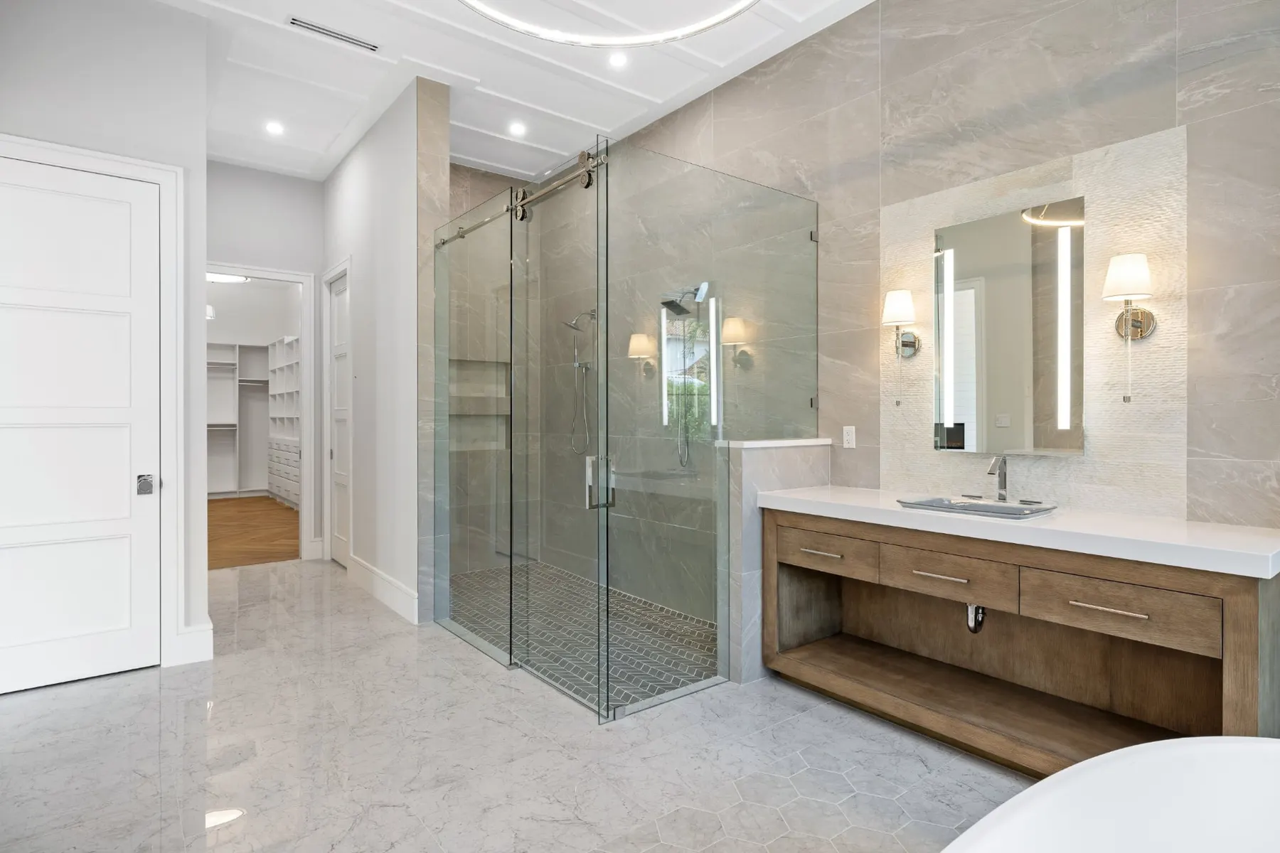Bath with a glass-enclosed shower, a wood vanity, a lit mirror and sconces, and a walk-in closet beyond.