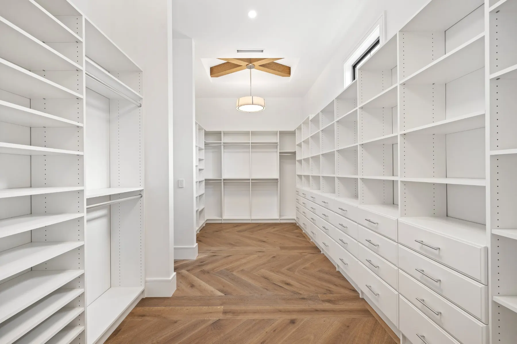 Walk-in closet with white shelving, drawers, hanging rods, a herringbone floor and a wood beam accent in the ceiling.