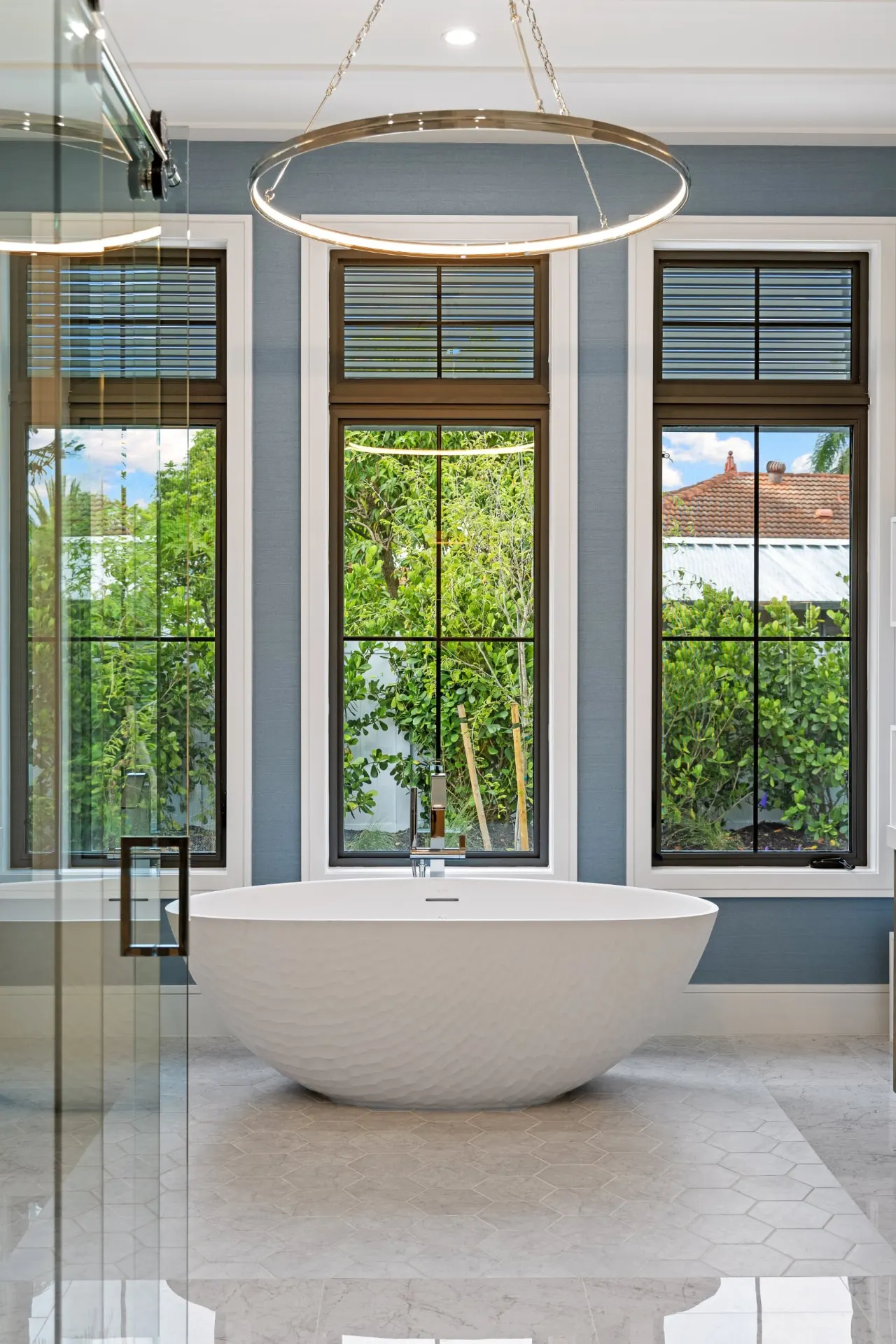Portrait view of the freestanding tub with a ring chandelier and three tall windows.