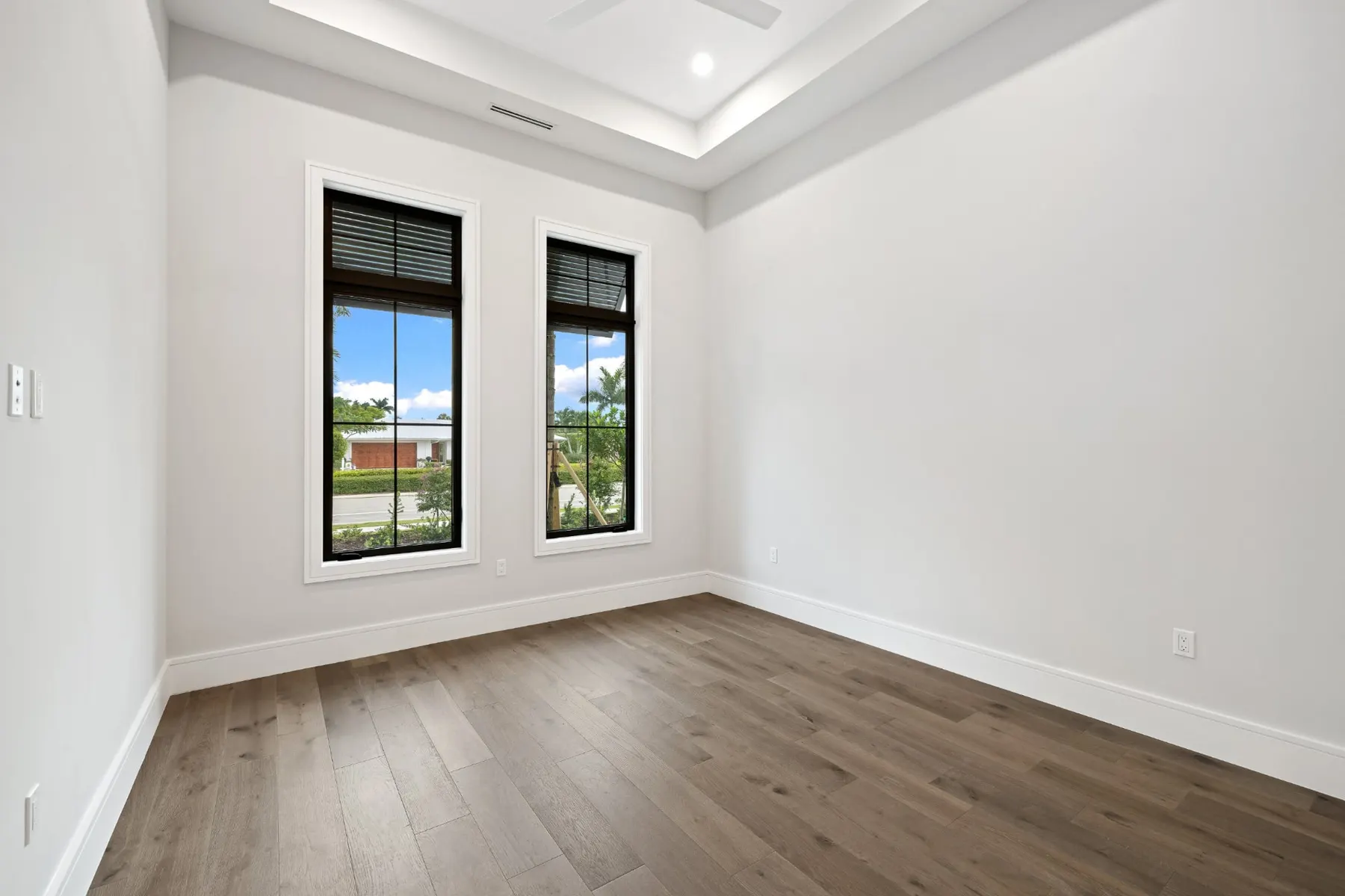 Bedroom with a tray ceiling, two tall black-framed windows and a plank wood floor.