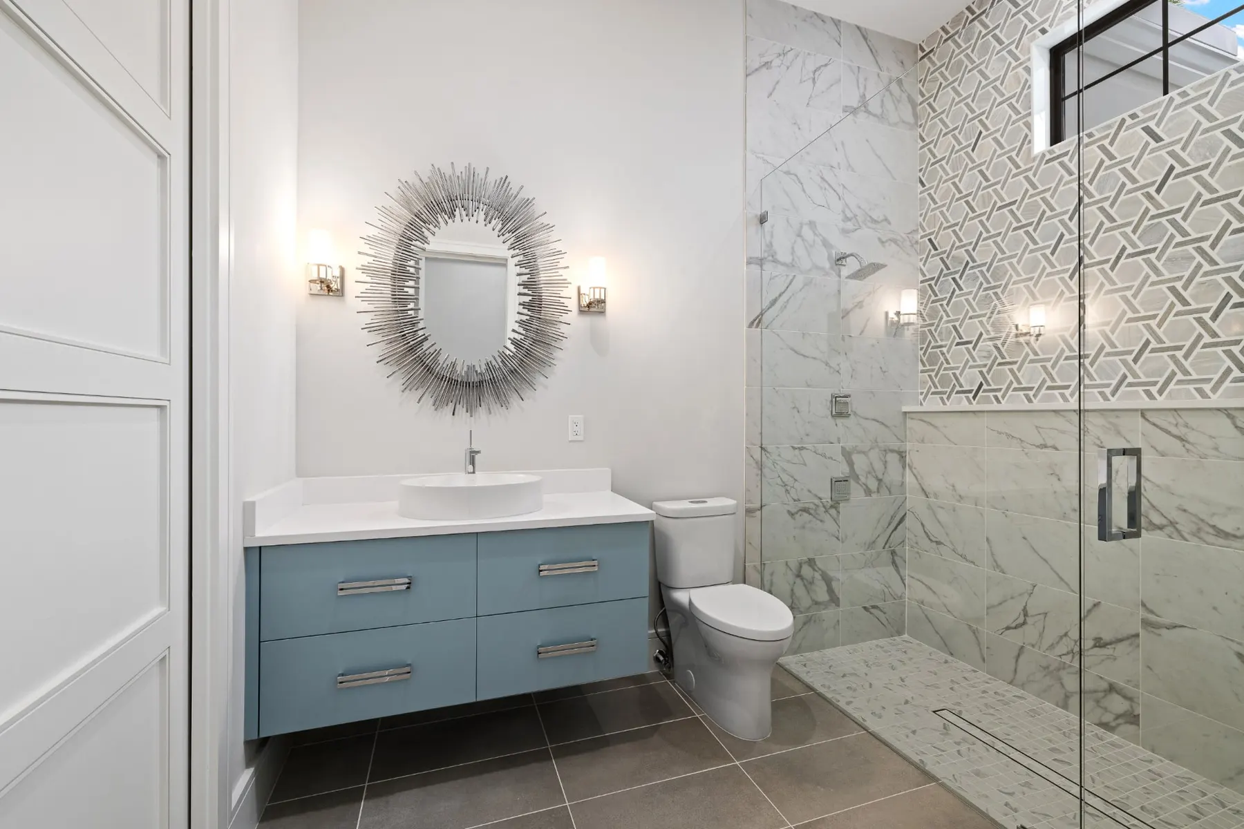 Bath with a blue floating vanity, a sunburst mirror and a marble-look walk-in shower.