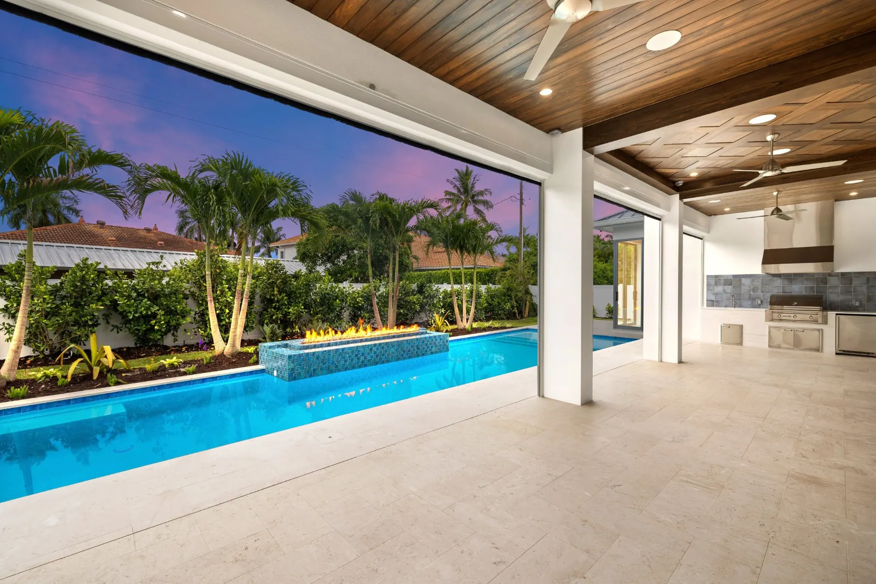 View from the covered lanai across the pool and fire feature at dusk, with the outdoor kitchen at right.