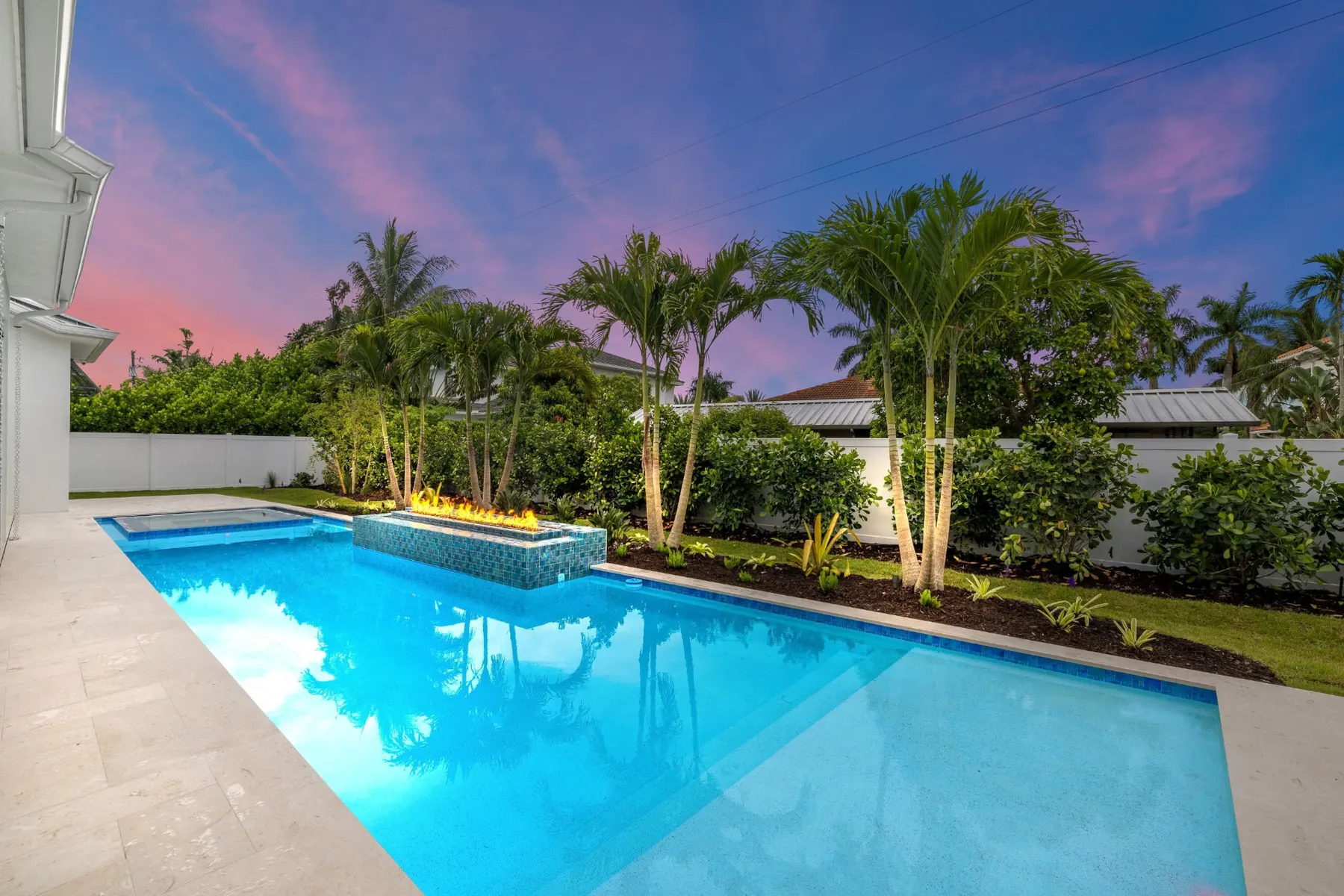 Lap pool with a spa and raised fire feature at dusk, framed by palms and landscaping.