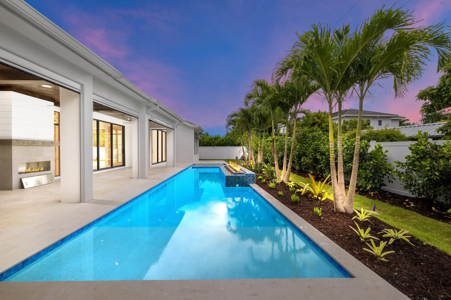 Long lap pool beside the covered lanai at dusk, with a fire feature and a lanai fireplace.