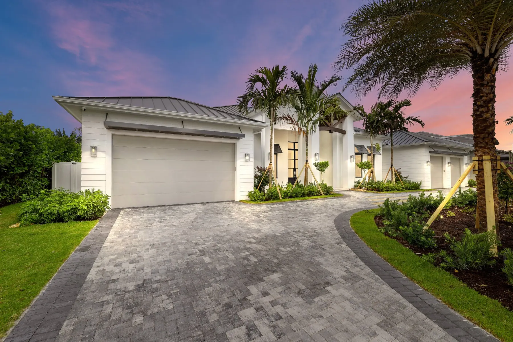 Front of the home at dusk from the paver driveway, with a two-car garage, a standing-seam metal roof and the lit timber-trussed entry.