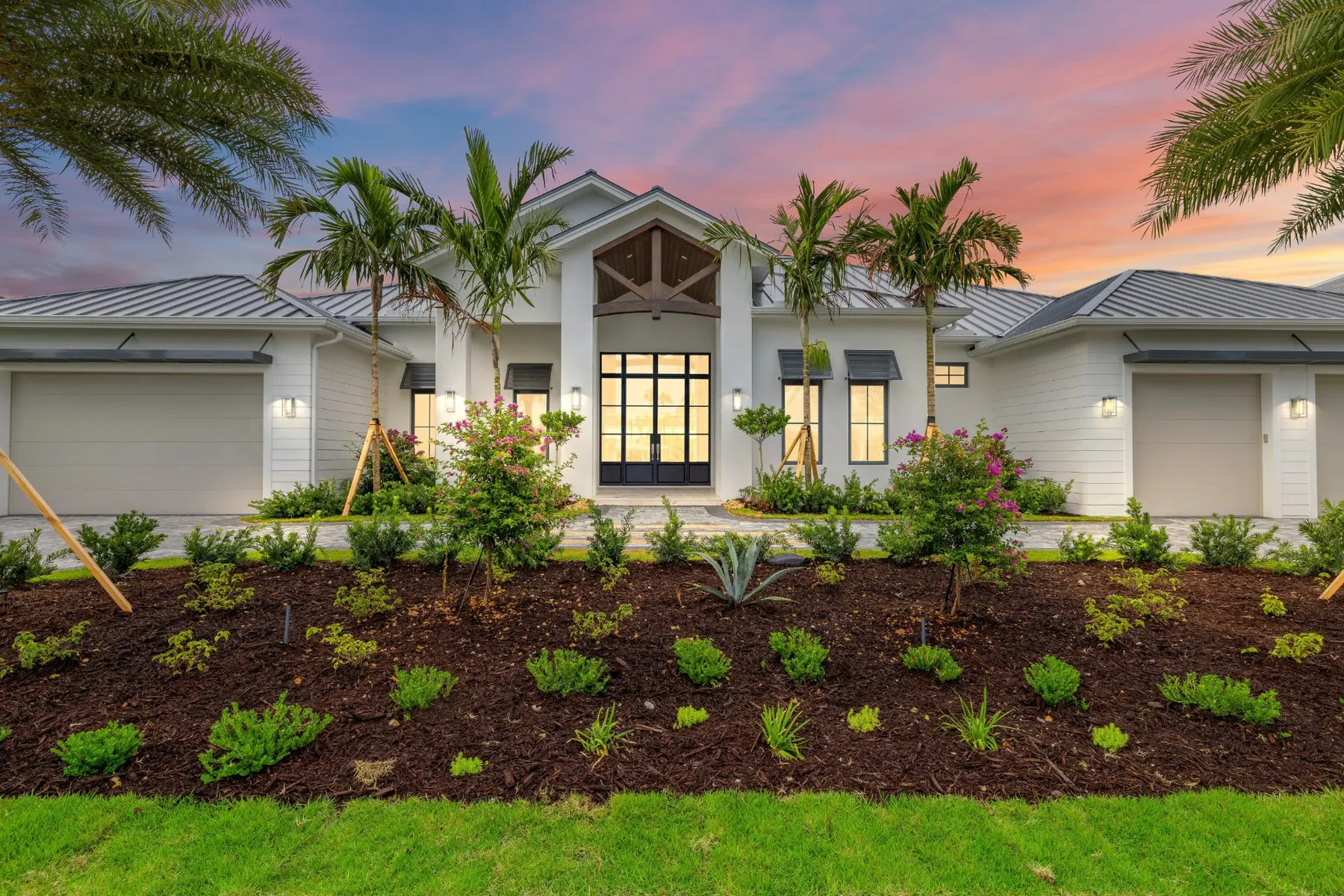 Front of The Moorings Residence at dusk, a white single-story home with a metal roof, a timber-trussed entry gable, black-framed glass double doors and a landscaped front bed.