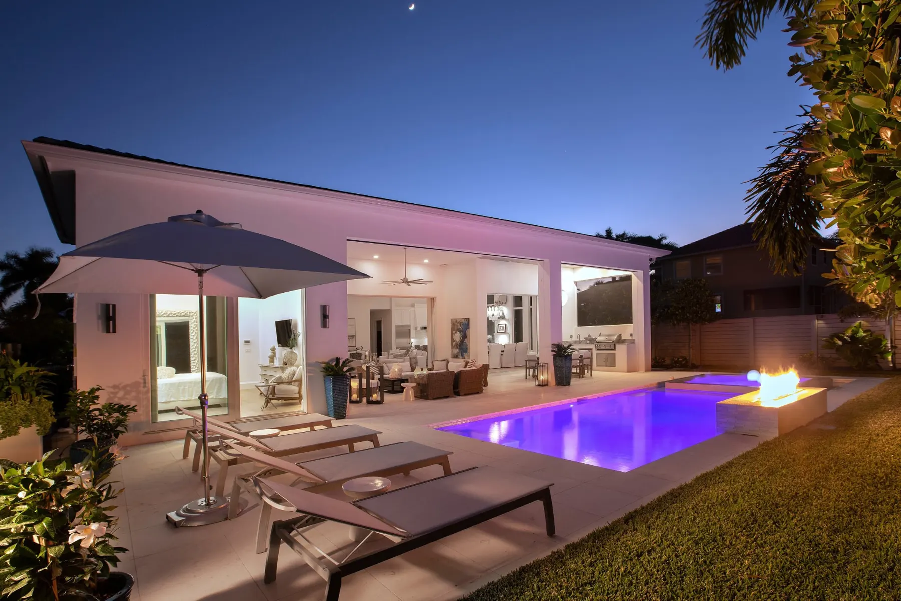 Covered lanai and pool terrace at dusk with lounge chairs, an umbrella and a fire feature beside the pool.
