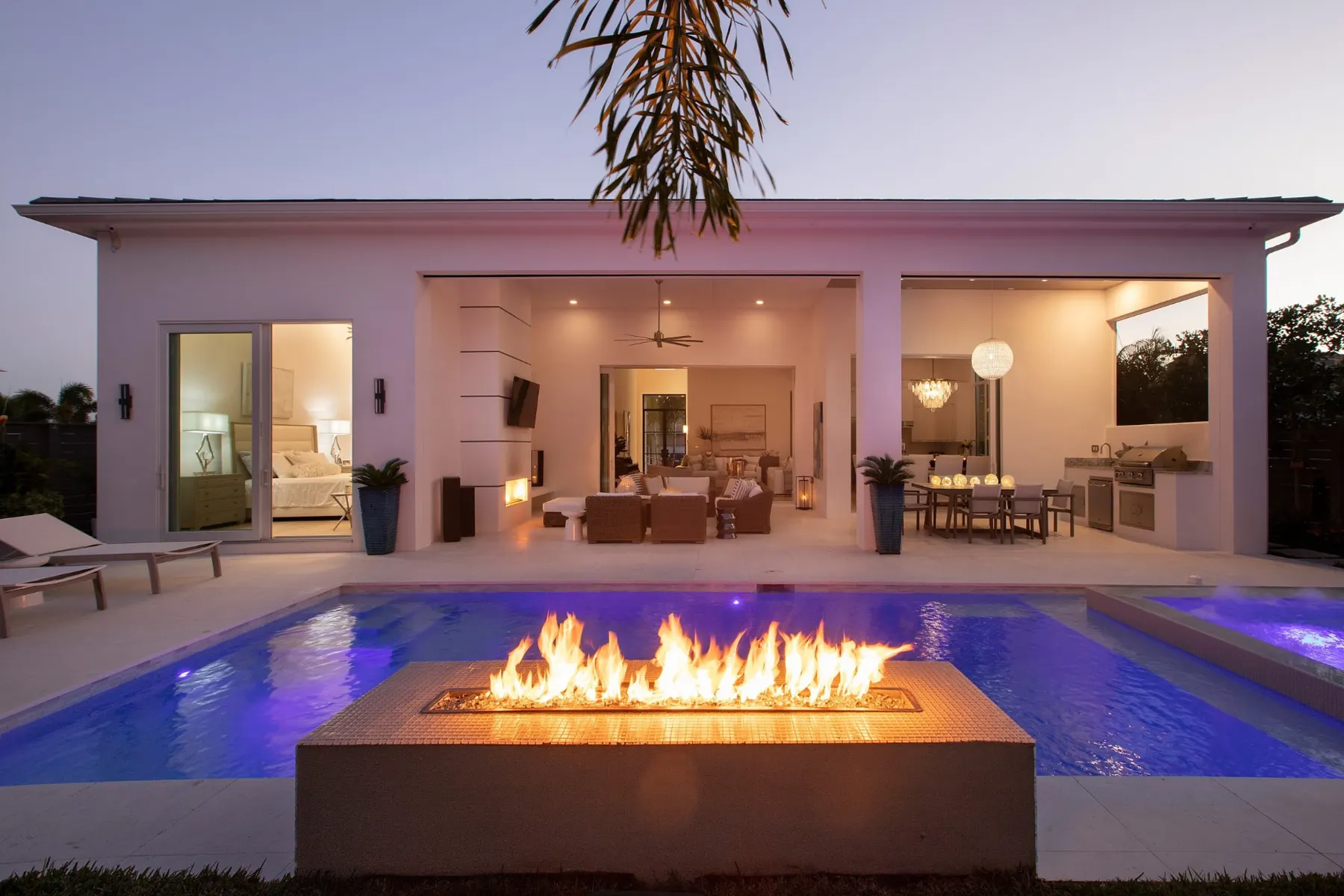 Pool fire feature in the foreground at dusk with the lit covered lanai and primary bedroom doors behind it.
