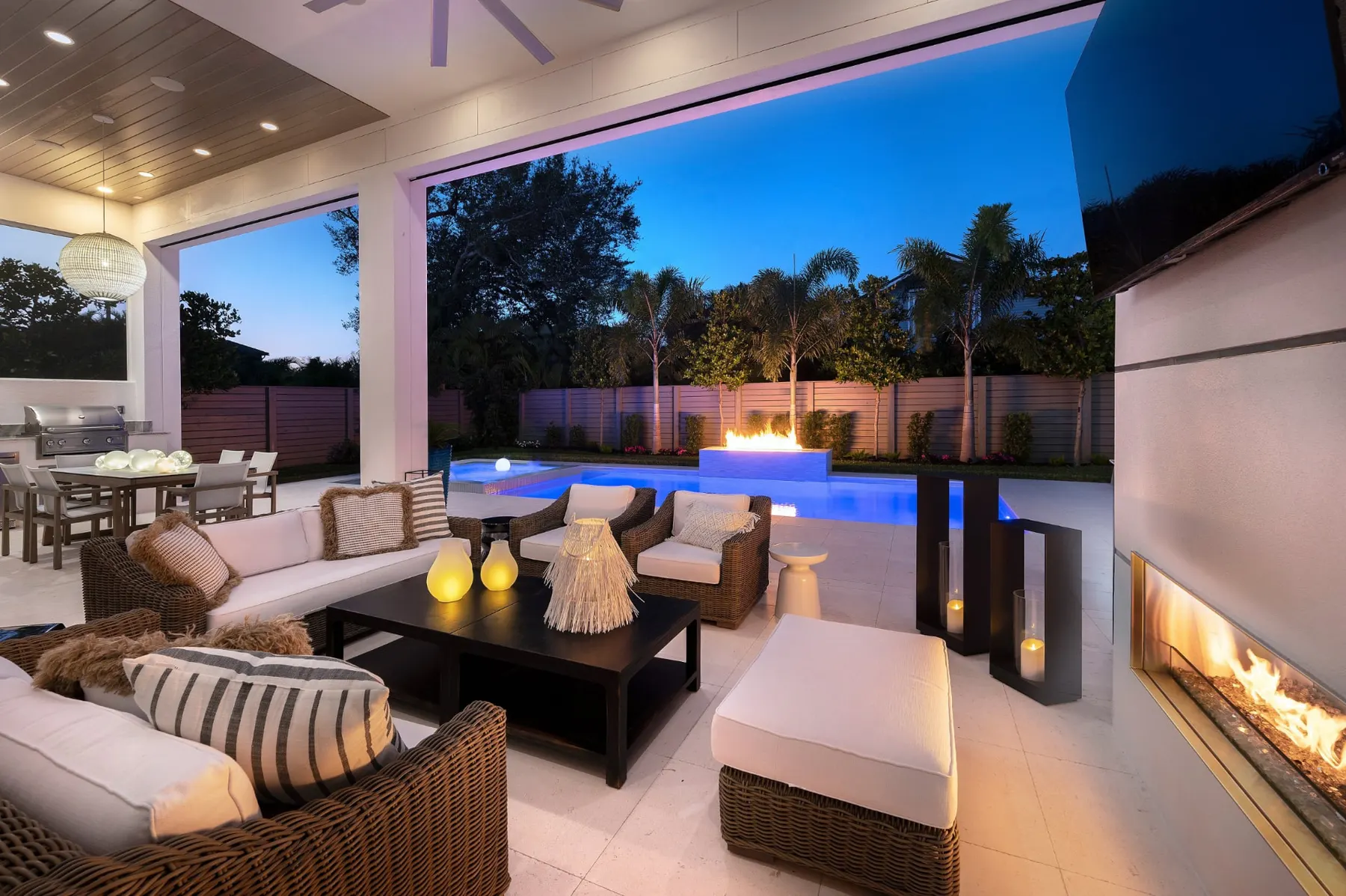 Covered outdoor living area at dusk with wicker seating, a linear fireplace and the lit pool beyond.