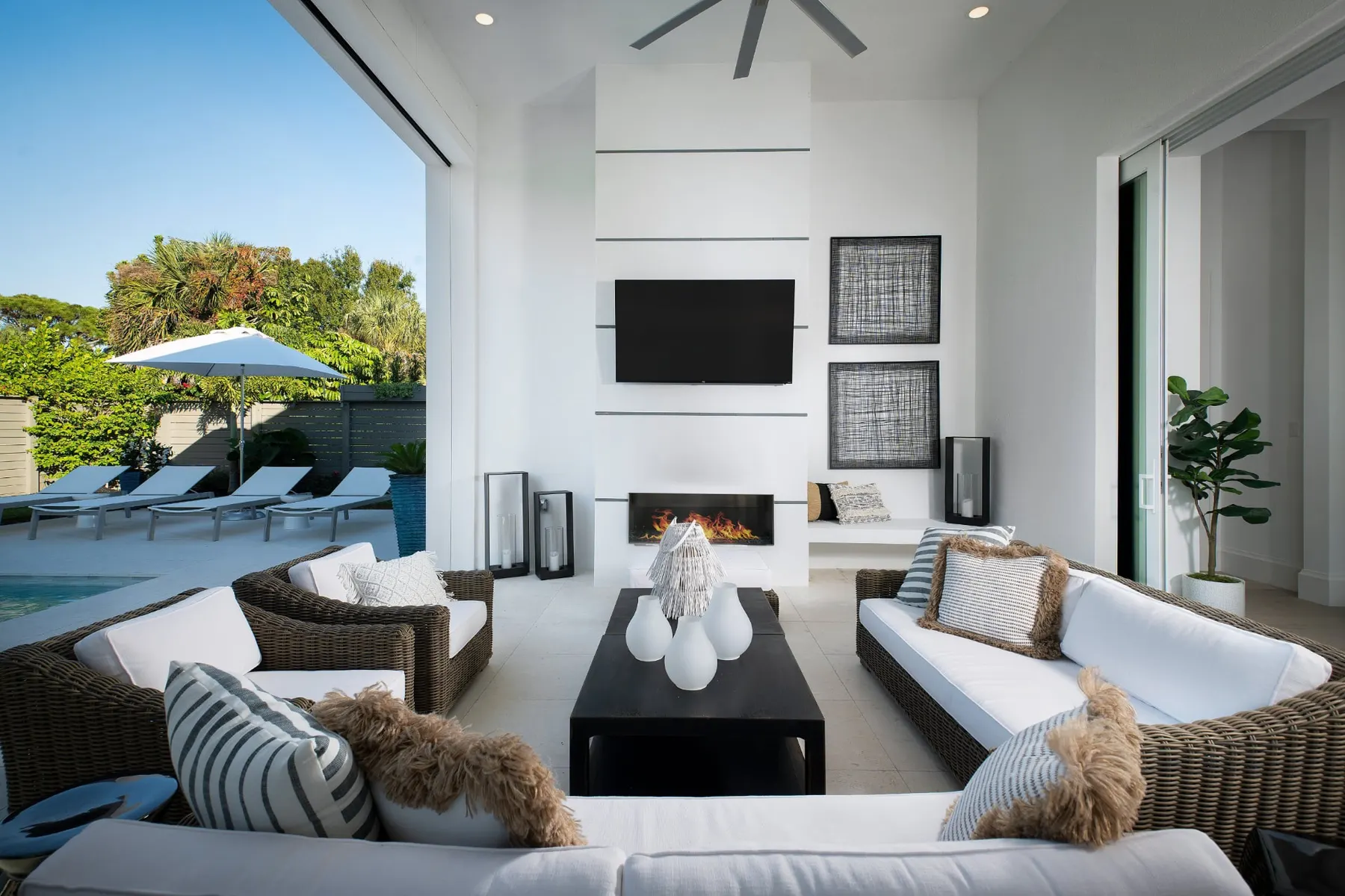 Covered lanai seating area by day with a linear fireplace, a wall-mounted television and lounge chairs by the pool.