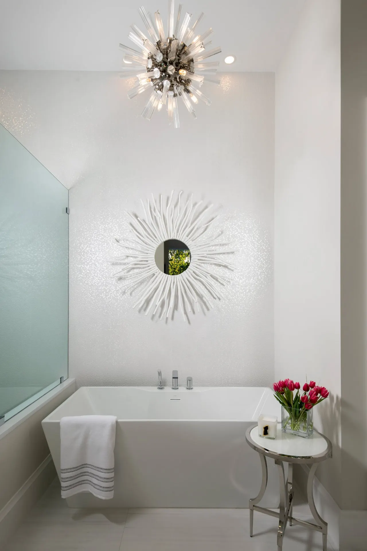 Freestanding tub against a mosaic tile wall with a starburst mirror and a chandelier above.