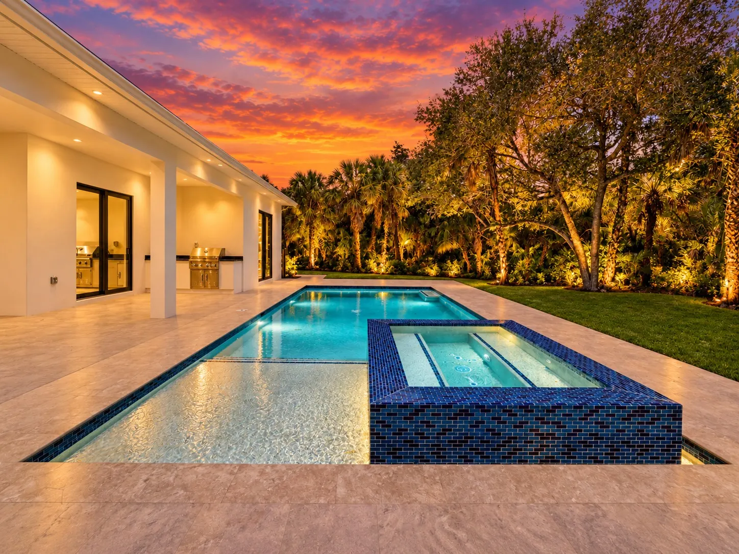Rectangular pool with a blue glass-tile spa and covered lanai with outdoor kitchen at dusk.