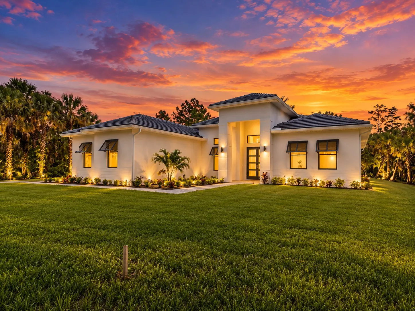 Front of The Estates Contemporary at dusk, a single-story white stucco home with a tall entry, dark tile roof and bahama-style shutters.