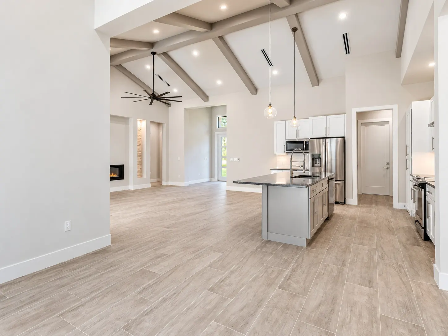Open great room and kitchen with exposed ceiling beams, a stone-top island and white shaker cabinets.